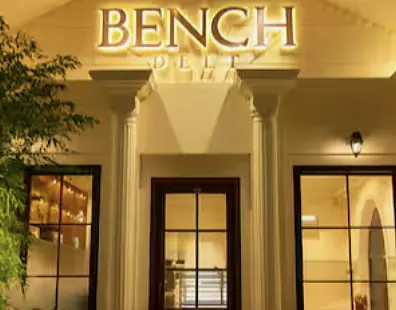 The lit portico of Bench Deli at dusk, seen through the greenery